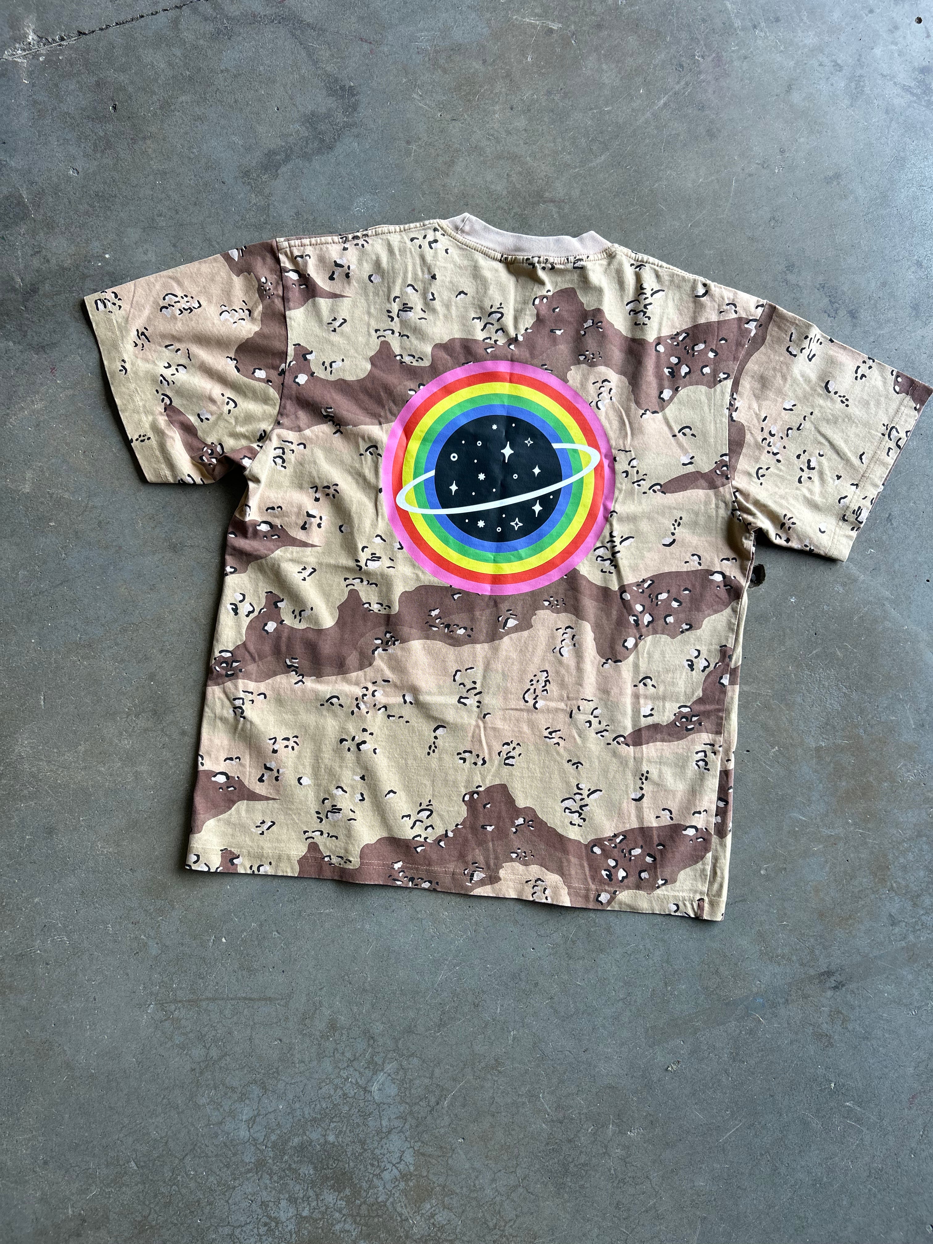 A short-sleeve t-shirt with a brown and beige desert camouflage pattern, featuring a large circular graphic on the chest with a rainbow ring around a black space planet, laid flat on a gray concrete floor.