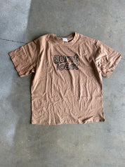 A brown short-sleeve t-shirt with 'Burnt Toast' printed in black outline text on the chest, laid flat on a gray concrete floor.