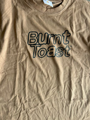 A close-up of a tan crewneck t-shirt with 'Burnt Toast' printed in a dark outline font across the chest.