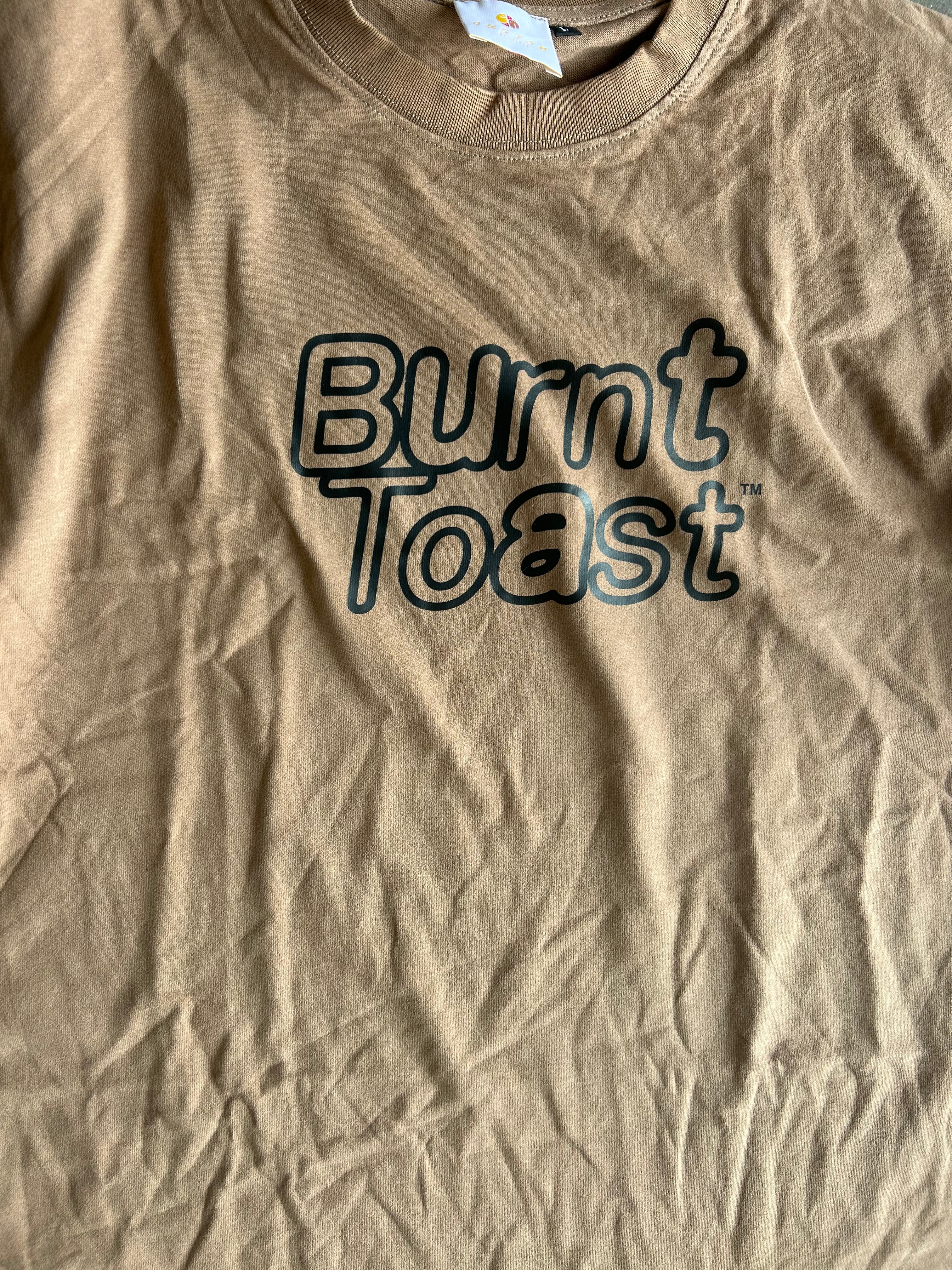 A close-up of a tan crewneck t-shirt with 'Burnt Toast' printed in a dark outline font across the chest.