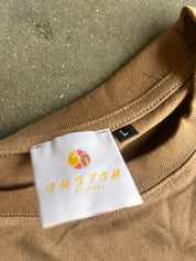 Close-up of a brown ribbed knit garment neck with a white sewn-in label reading CUSTOM and a small black size tag marked L, placed on a speckled gray surface.