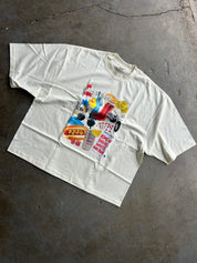 An oversized off-white t-shirt laid flat on a textured gray concrete floor, featuring a colorful graphic print on the front with various retro-themed illustrations and text.