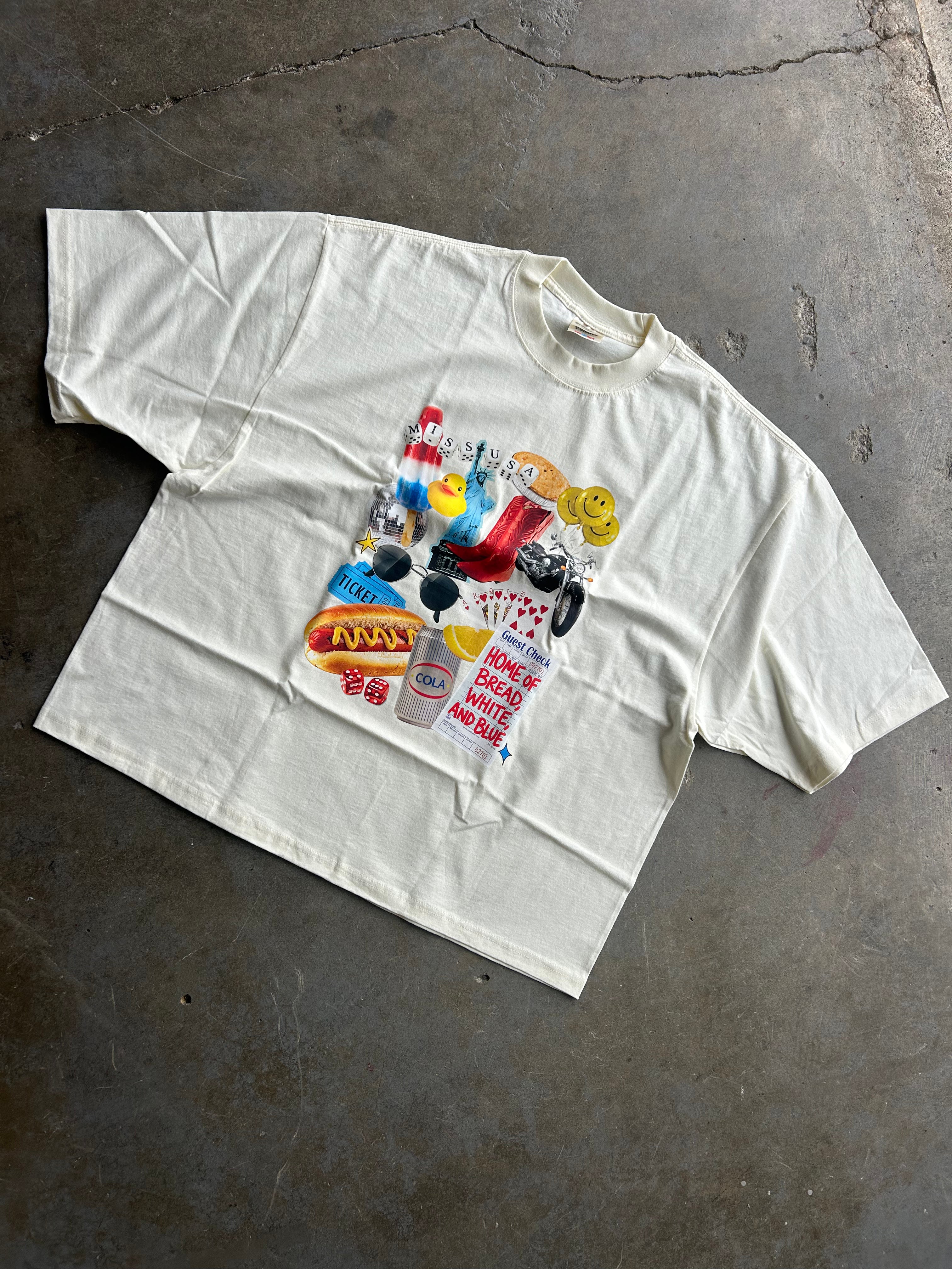 An oversized off-white t-shirt laid flat on a textured gray concrete floor, featuring a colorful graphic print on the front with various retro-themed illustrations and text.