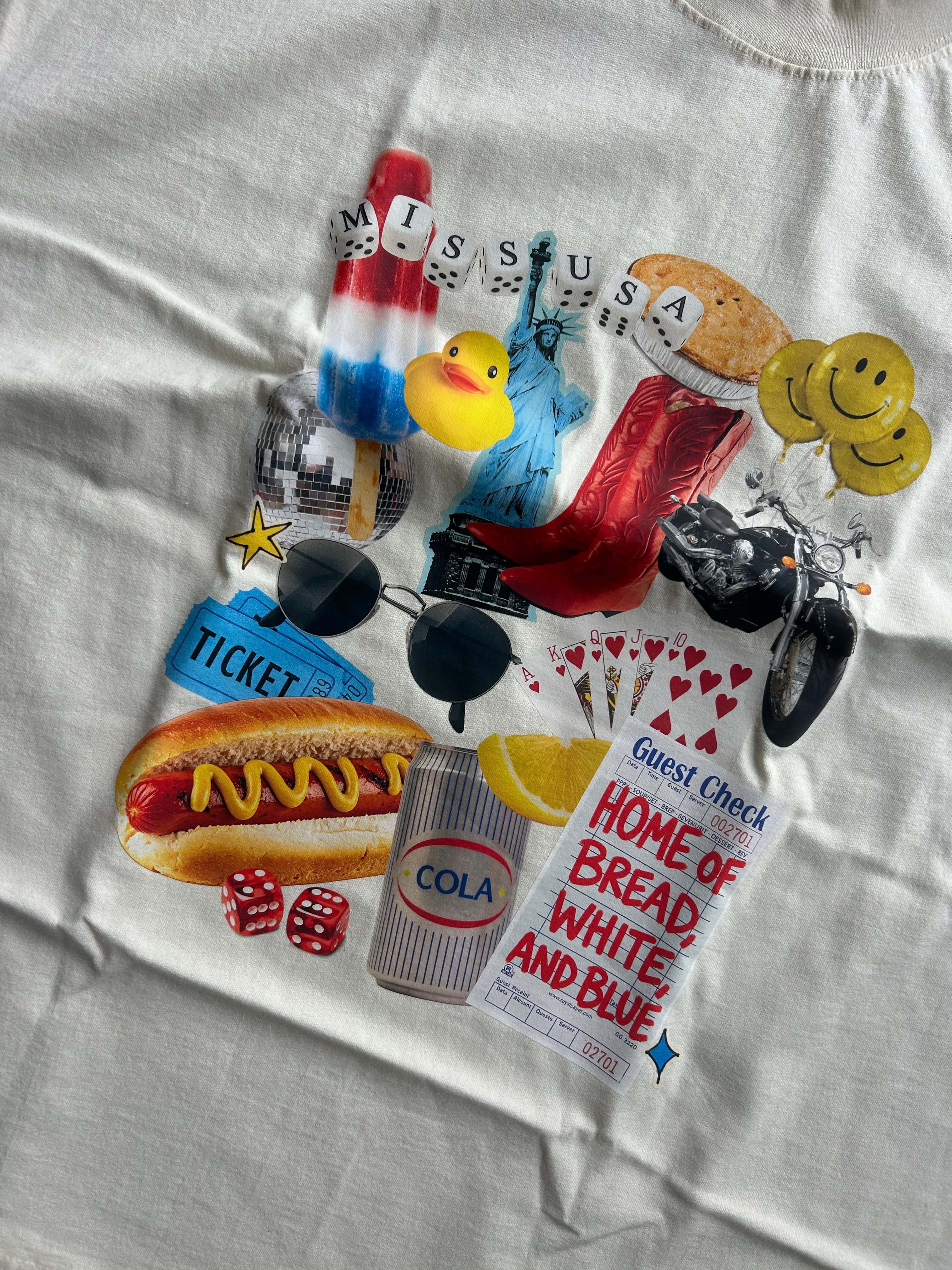 Close up of an oversized off-white t-shirt laid flat on a textured gray concrete floor, featuring a colorful graphic print on the front with various retro-themed illustrations and text.