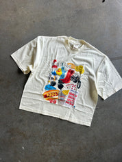 An oversized cream-colored graphic t-shirt laid flat on a gray concrete floor. The shirt features a large, colorful collage print on the front with retro cartoon characters, playing cards, a red high-top sneaker, and text 