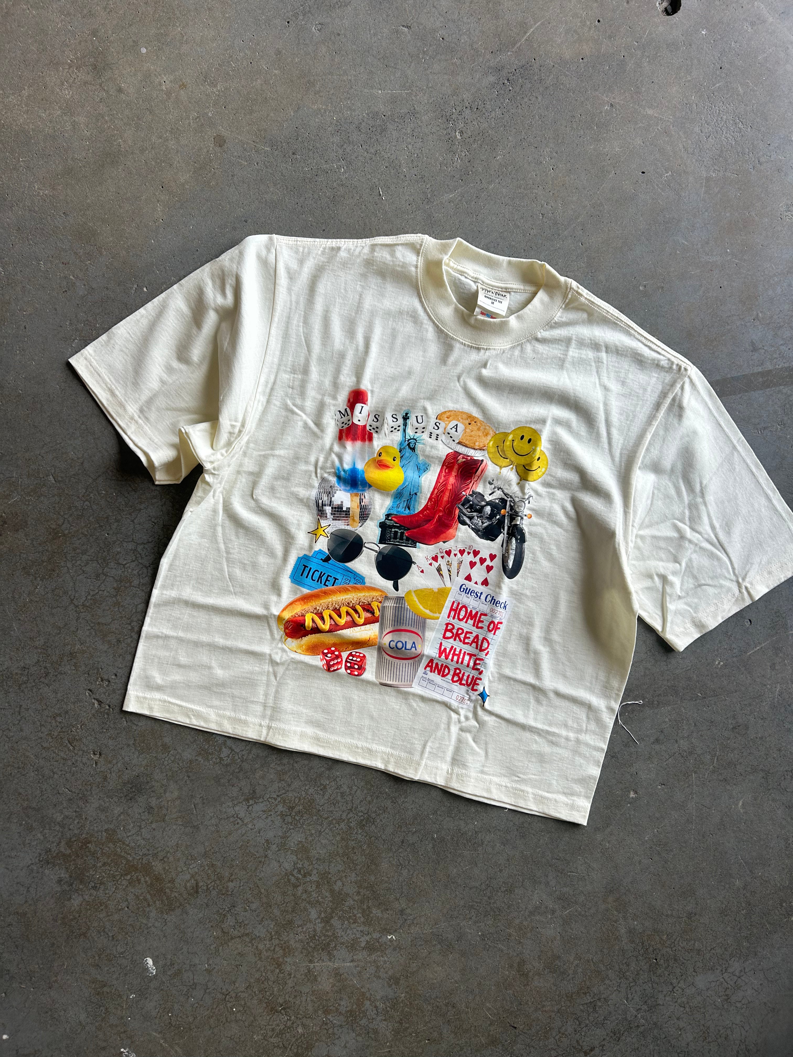 An oversized cream-colored graphic t-shirt laid flat on a gray concrete floor. The shirt features a large, colorful collage print on the front with retro cartoon characters, playing cards, a red high-top sneaker, and text 