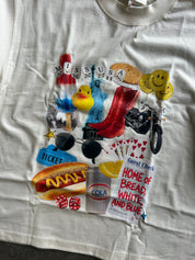 A white graphic t-shirt laid flat, featuring a colorful collage print with various pop-culture and retro-themed illustrations including a hot dog, yellow rubber duck, red high heels, a motorcycle, smiley faces, and text reading 'Miss USA' and 'Home of Bread, White, and Blue'.