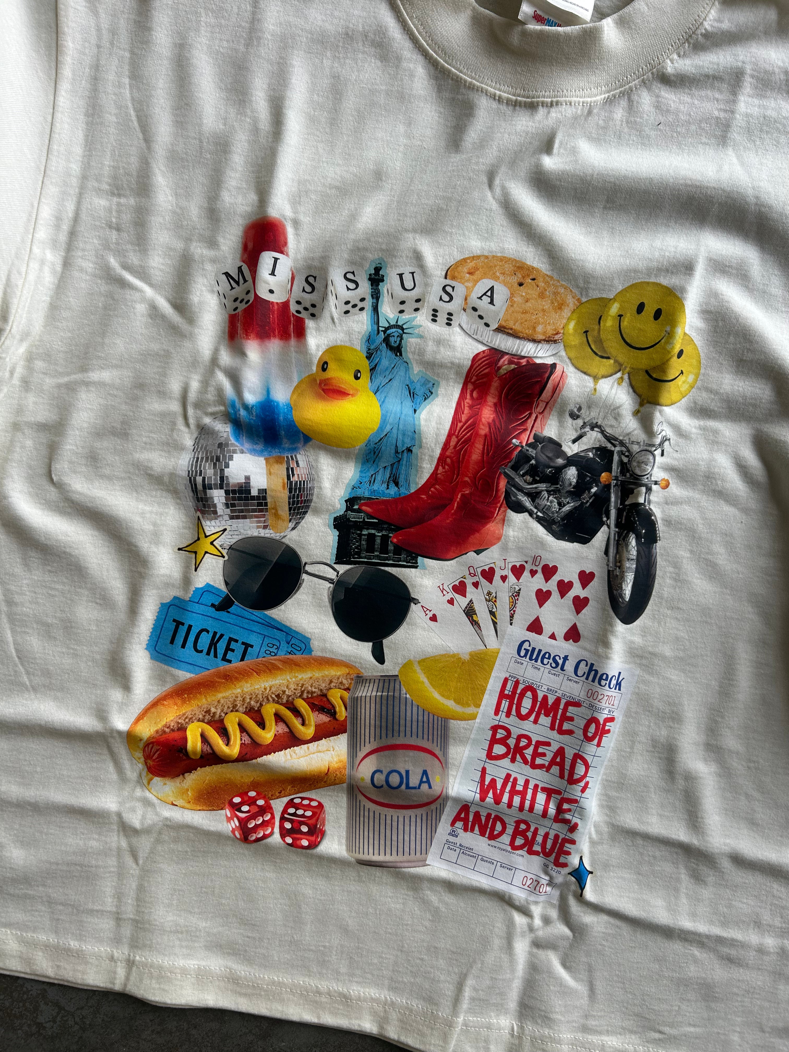 A white graphic t-shirt laid flat, featuring a colorful collage print with various pop-culture and retro-themed illustrations including a hot dog, yellow rubber duck, red high heels, a motorcycle, smiley faces, and text reading 'Miss USA' and 'Home of Bread, White, and Blue'.