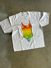 A white short-sleeve t-shirt laid flat on a concrete floor, featuring a colorful rainbow gradient graphic of a one-piece swimsuit patterned with yellow smiley faces.