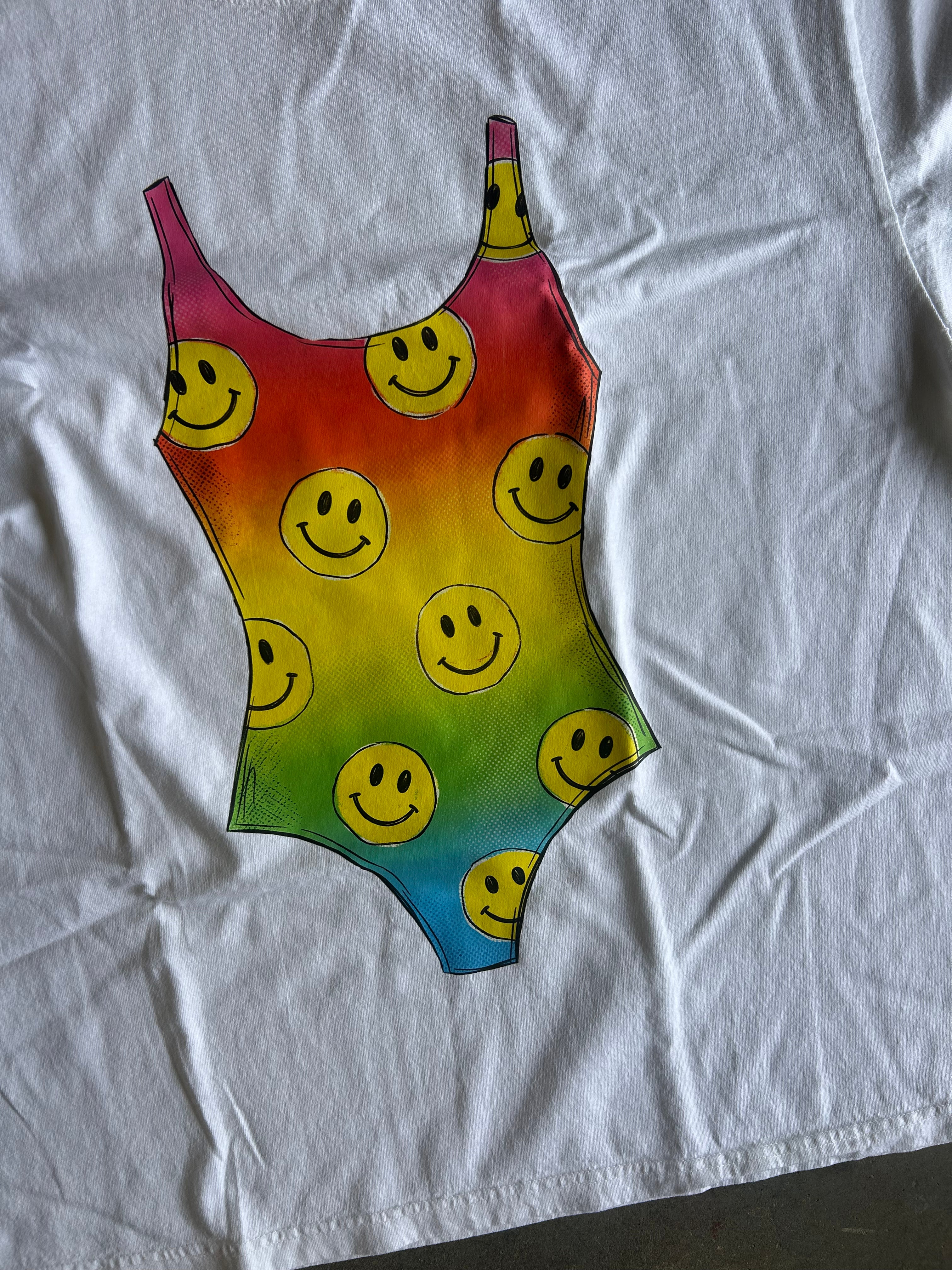 A white short-sleeve T-shirt laid flat, featuring a large graphic of a one-piece swimsuit with a rainbow gradient and a repeating yellow smiley face pattern.