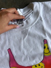 A close-up of a white heavyweight t-shirt laid flat, showing a hand pointing to the inner neck label which reads 'FAMOUS HEAVYWEIGHT T-SHIRTS'. The t-shirt features a colorful graphic of a banana split at the bottom.