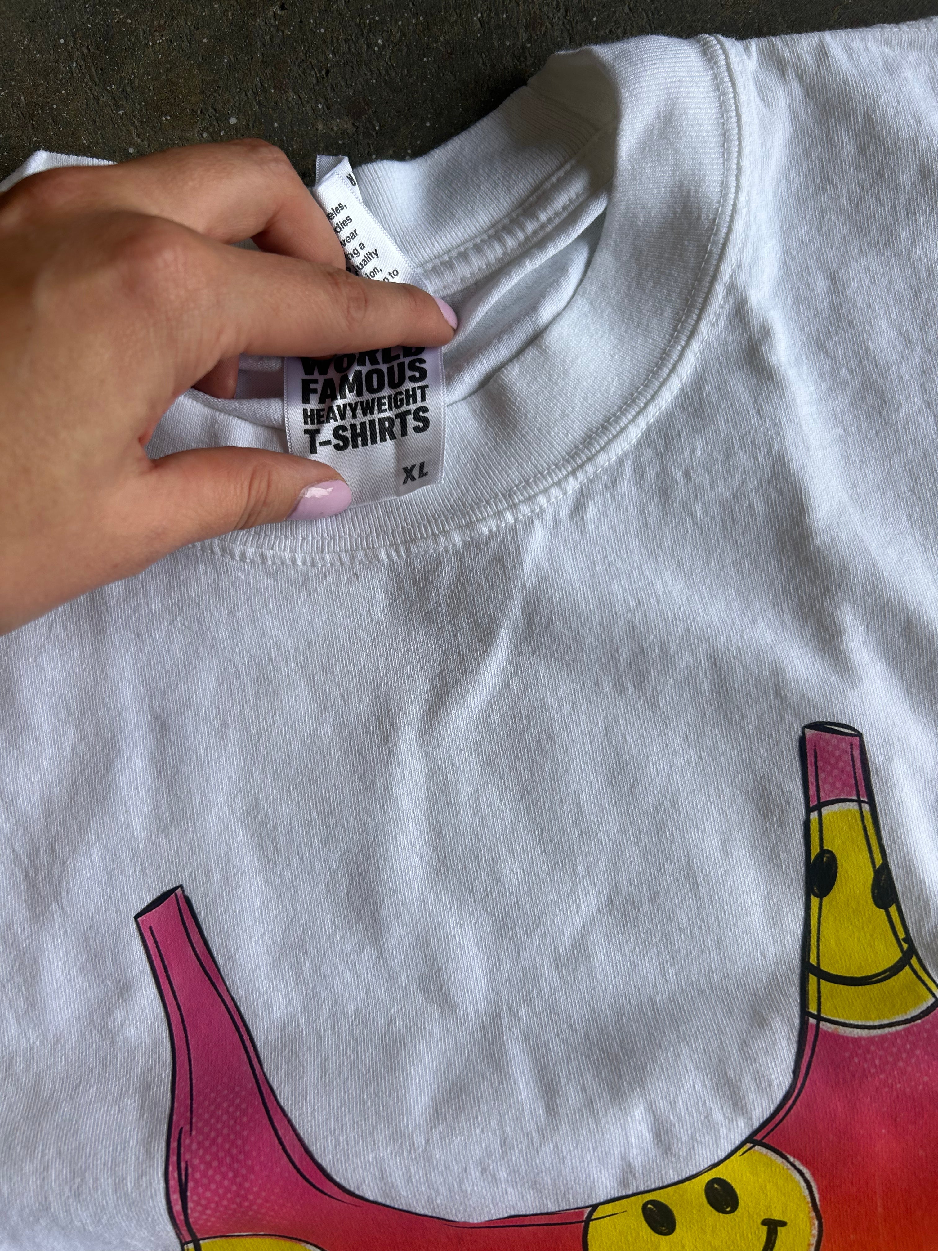 A close-up of a white heavyweight t-shirt laid flat, showing a hand pointing to the inner neck label which reads 'FAMOUS HEAVYWEIGHT T-SHIRTS'. The t-shirt features a colorful graphic of a banana split at the bottom.