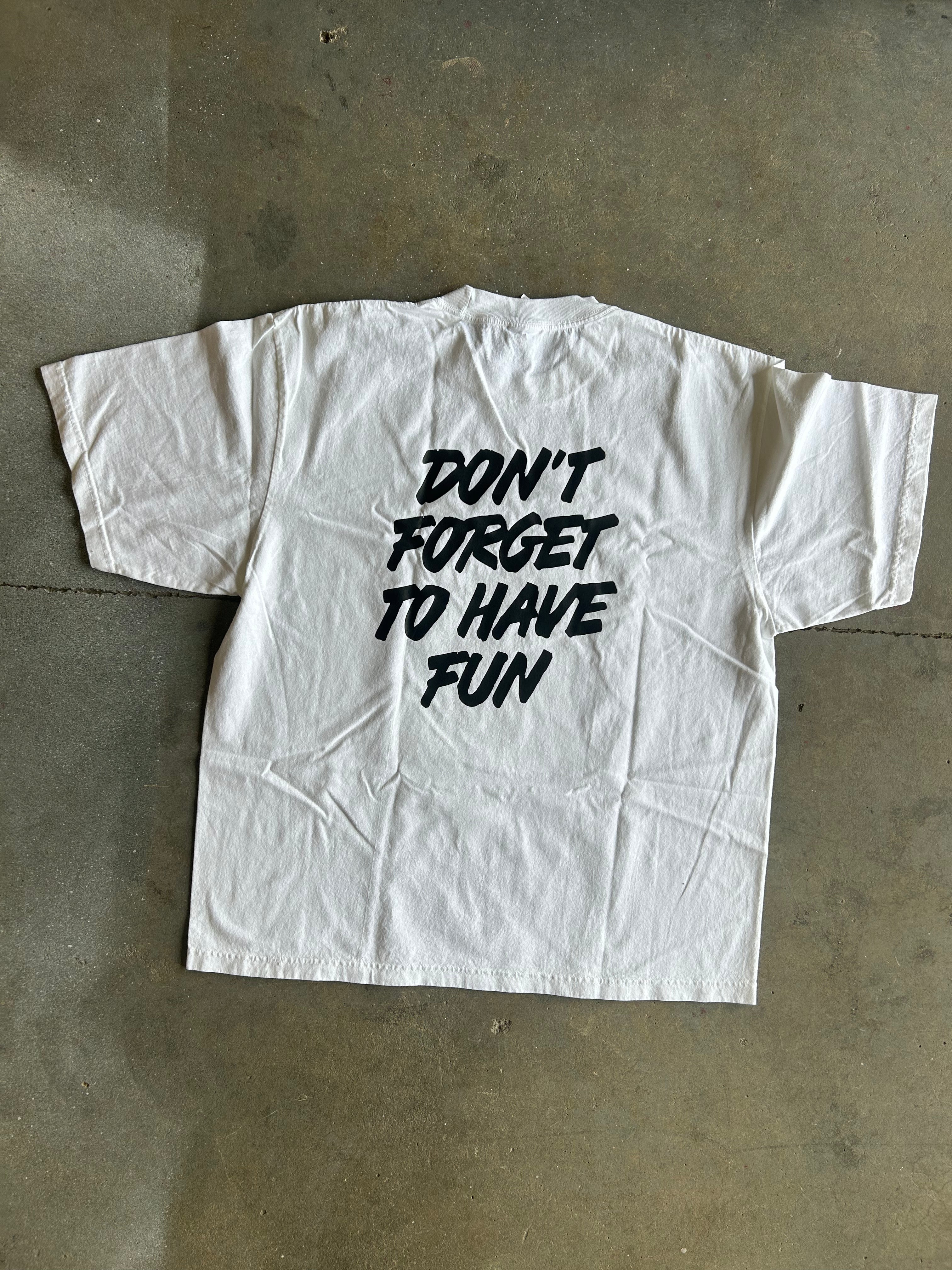 A white short-sleeve T-shirt laid flat on a concrete floor, featuring the phrase 'DON'T FORGET TO HAVE FUN' printed in black across the back.