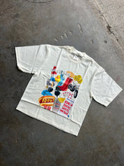 An off-white short-sleeve t-shirt with a colorful collage graphic on the front, laid flat on a textured gray concrete floor.