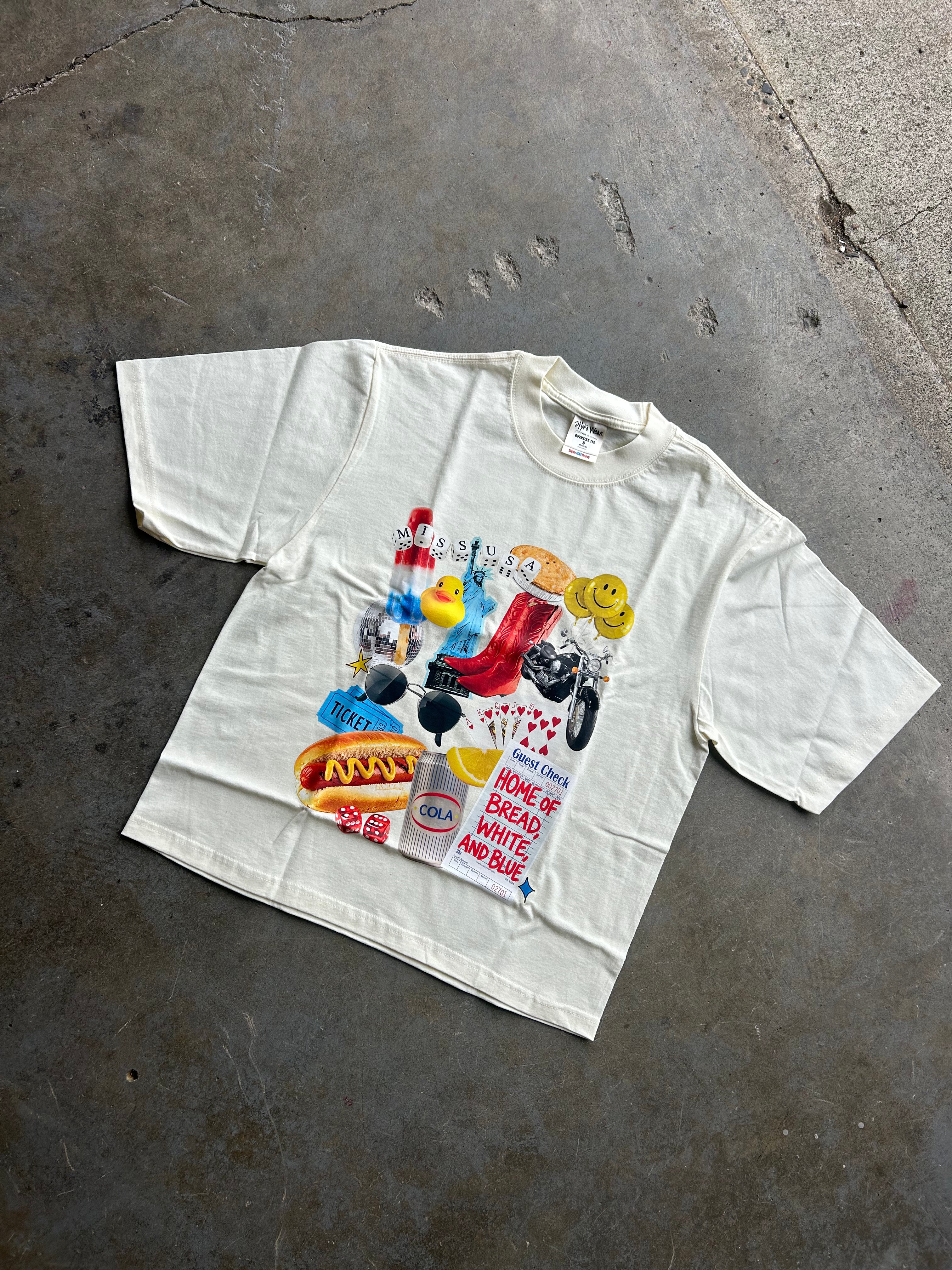 An off-white short-sleeve t-shirt with a colorful collage graphic on the front, laid flat on a textured gray concrete floor.