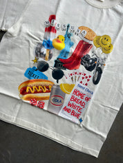 A white graphic T-shirt laid flat on a dark gray surface, featuring a colorful collage print with hot dogs, smiley faces, a motorcycle, a duck, dice, sunglasses, a ticket, and text including 'Miss USA' and 'Home of Bread, White, and Blue'.