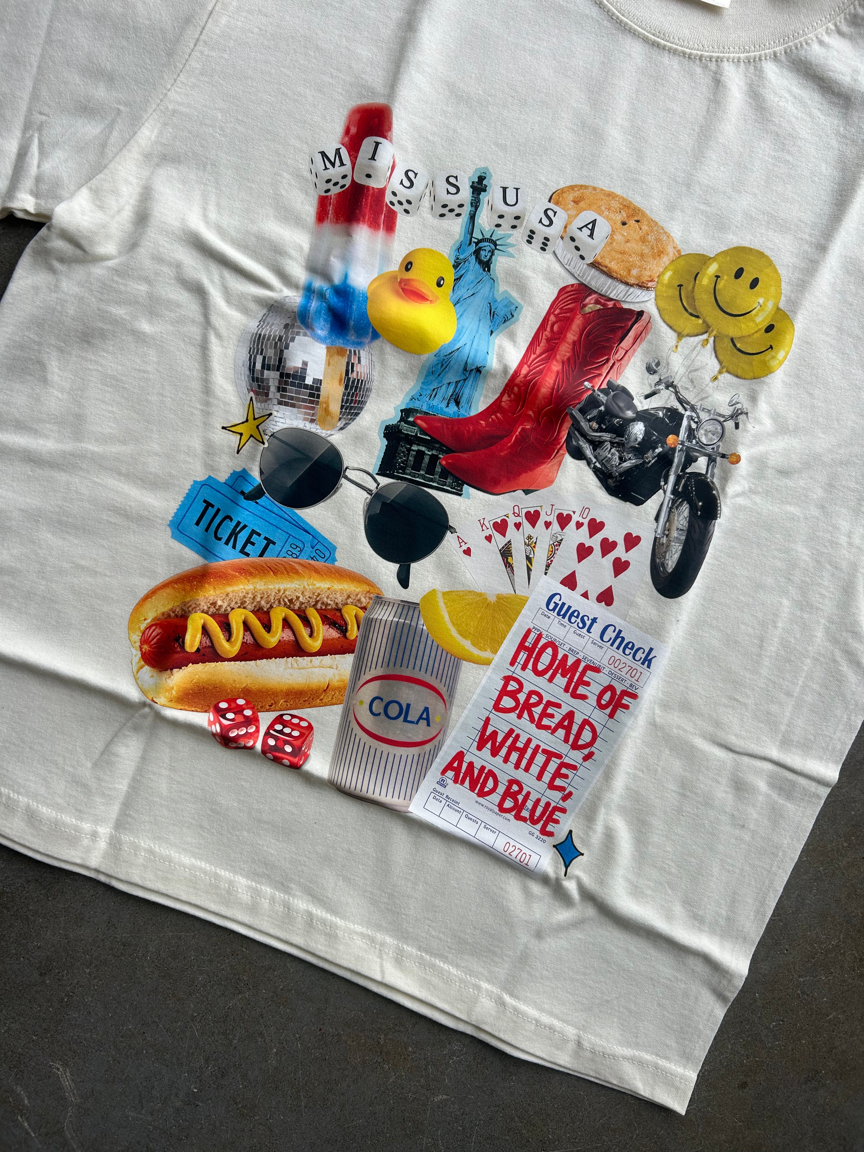 A white graphic T-shirt laid flat on a dark gray surface, featuring a colorful collage print with hot dogs, smiley faces, a motorcycle, a duck, dice, sunglasses, a ticket, and text including 'Miss USA' and 'Home of Bread, White, and Blue'.