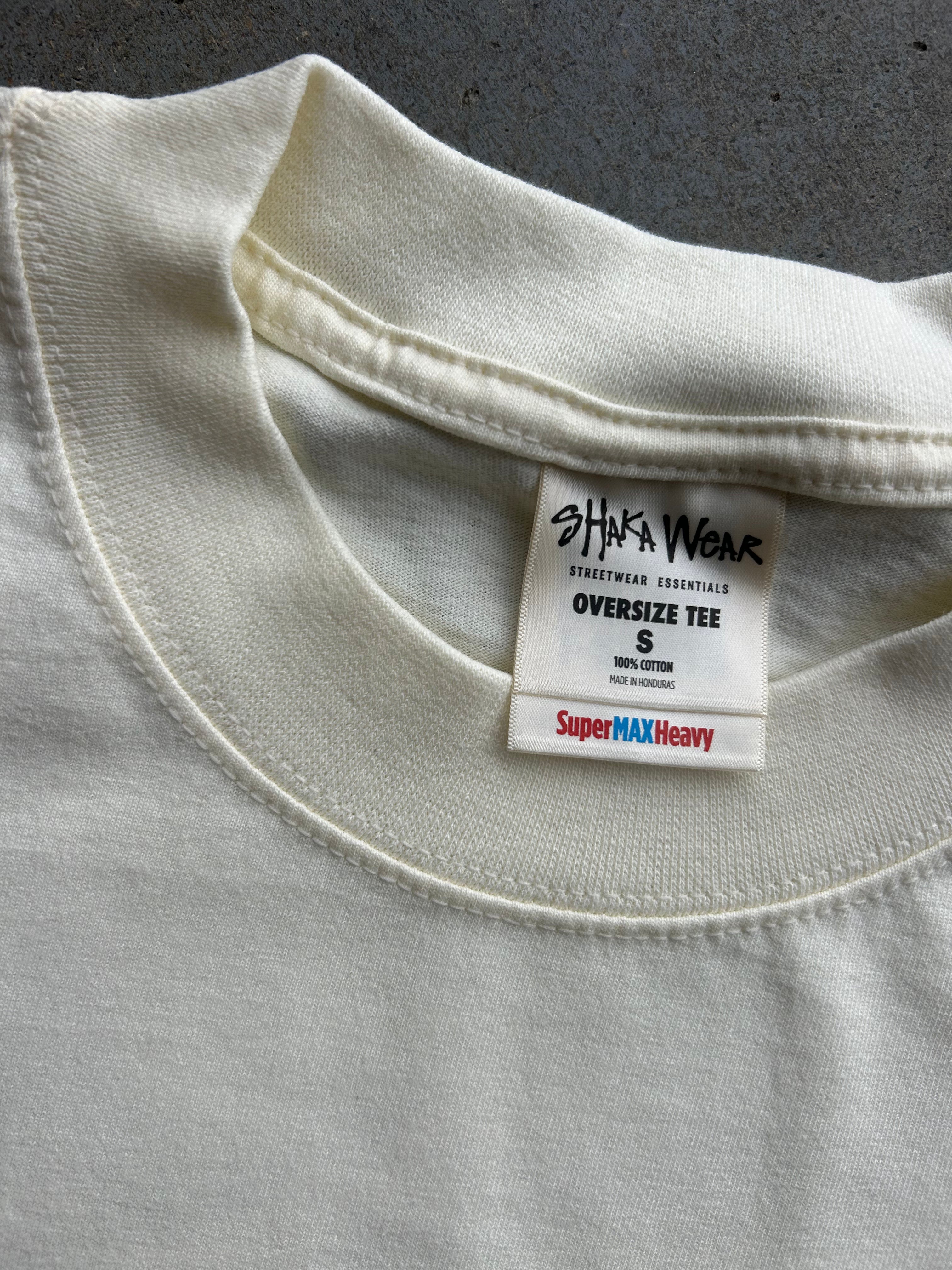 Close-up of a cream-colored oversized cotton t-shirt collar, showing the inner neck label for 'Shaka Wear' and 'SuperMAX Heavy' weight.