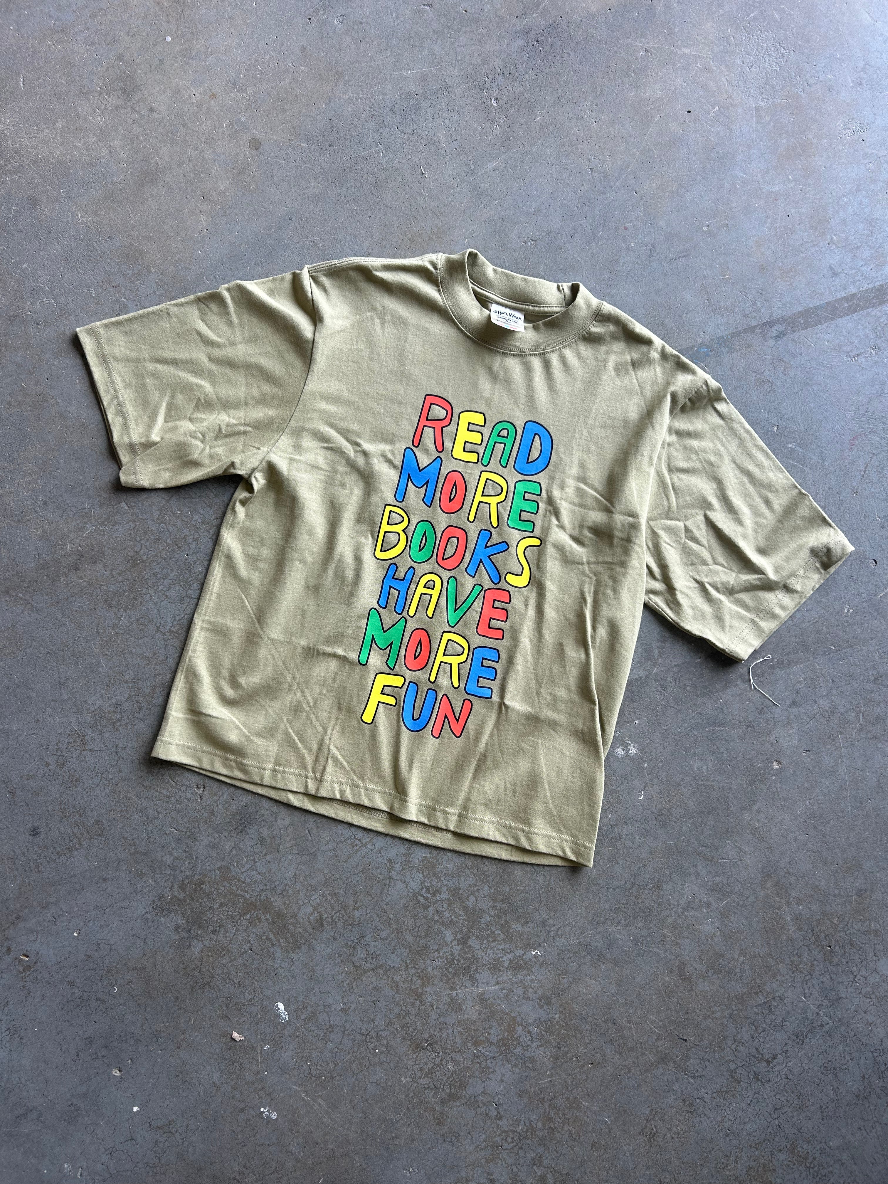 An oversized olive green t-shirt with a colorful retro-style graphic reading 'READ MORE BOOKS HAVE MORE FUN' in stacked, wavy letters, laid flat on a gray concrete floor