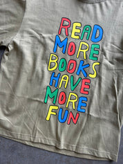 A close-up of a beige graphic t-shirt featuring the phrase 'READ MORE BOOKS HAVE MORE FUN' printed in large, colorful bubble letters outlined in black.