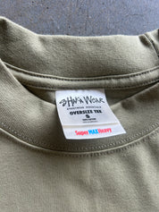 Close-up of the inside collar of an oversized olive green t-shirt, showing a white brand label for 'Shaka Wear' and a red and blue 'Super MAX Heavy' tag.