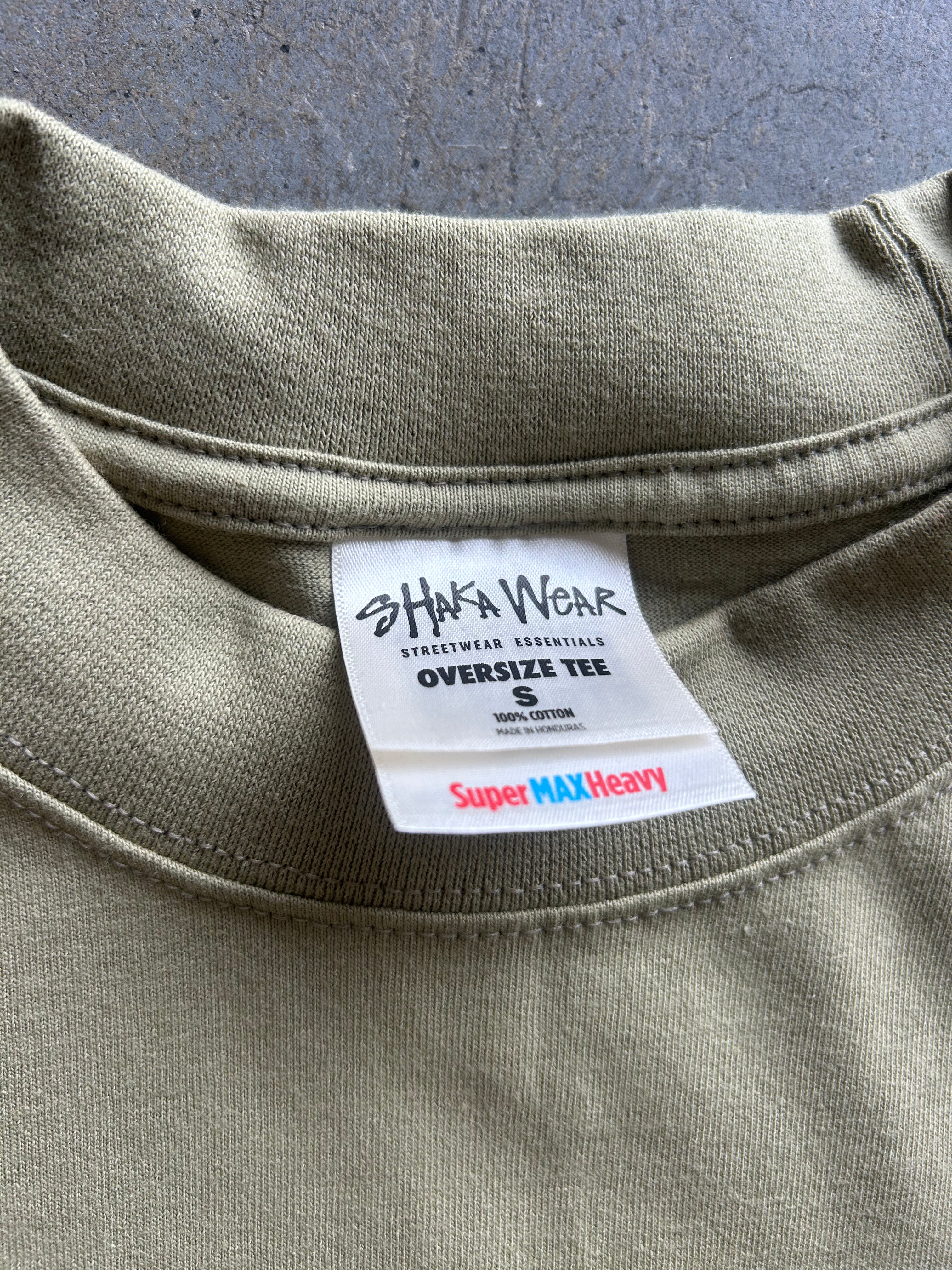 Close-up of the inside collar of an oversized olive green t-shirt, showing a white brand label for 'Shaka Wear' and a red and blue 'Super MAX Heavy' tag.