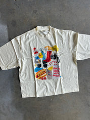 An oversized cream-colored short-sleeve t-shirt with a colorful graphic collage on the front, laid flat on a gray concrete floor.