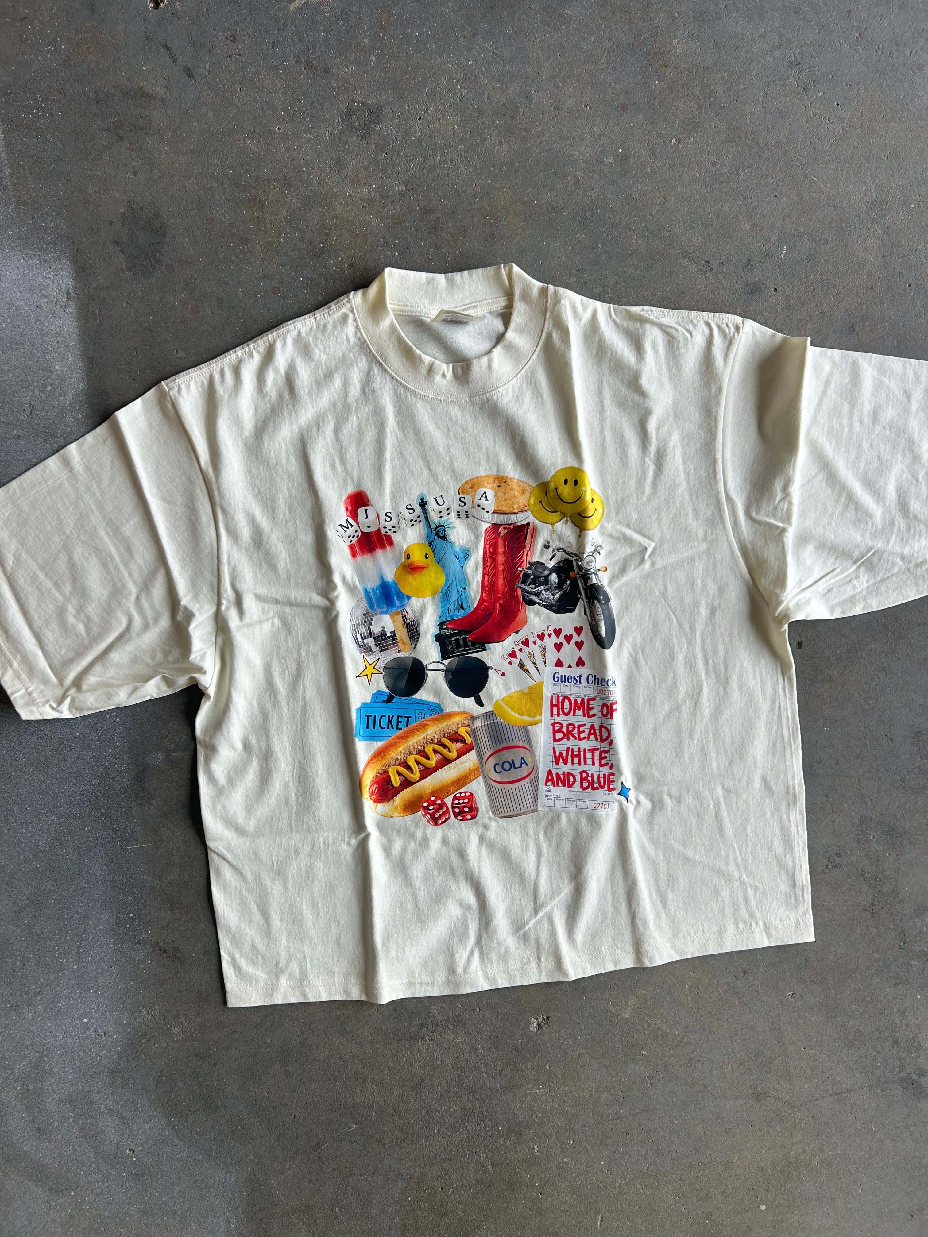 An oversized cream-colored short-sleeve t-shirt with a colorful graphic collage on the front, laid flat on a gray concrete floor.