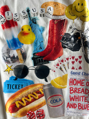 A white t-shirt featuring a colorful graphic collage of pop culture and retro elements, including a hot dog, cowboy boots, a yellow rubber duck, a Statue of Liberty, a motorcycle, sunglasses, a hot dog, a soda can, playing cards, dice, and a ticket.