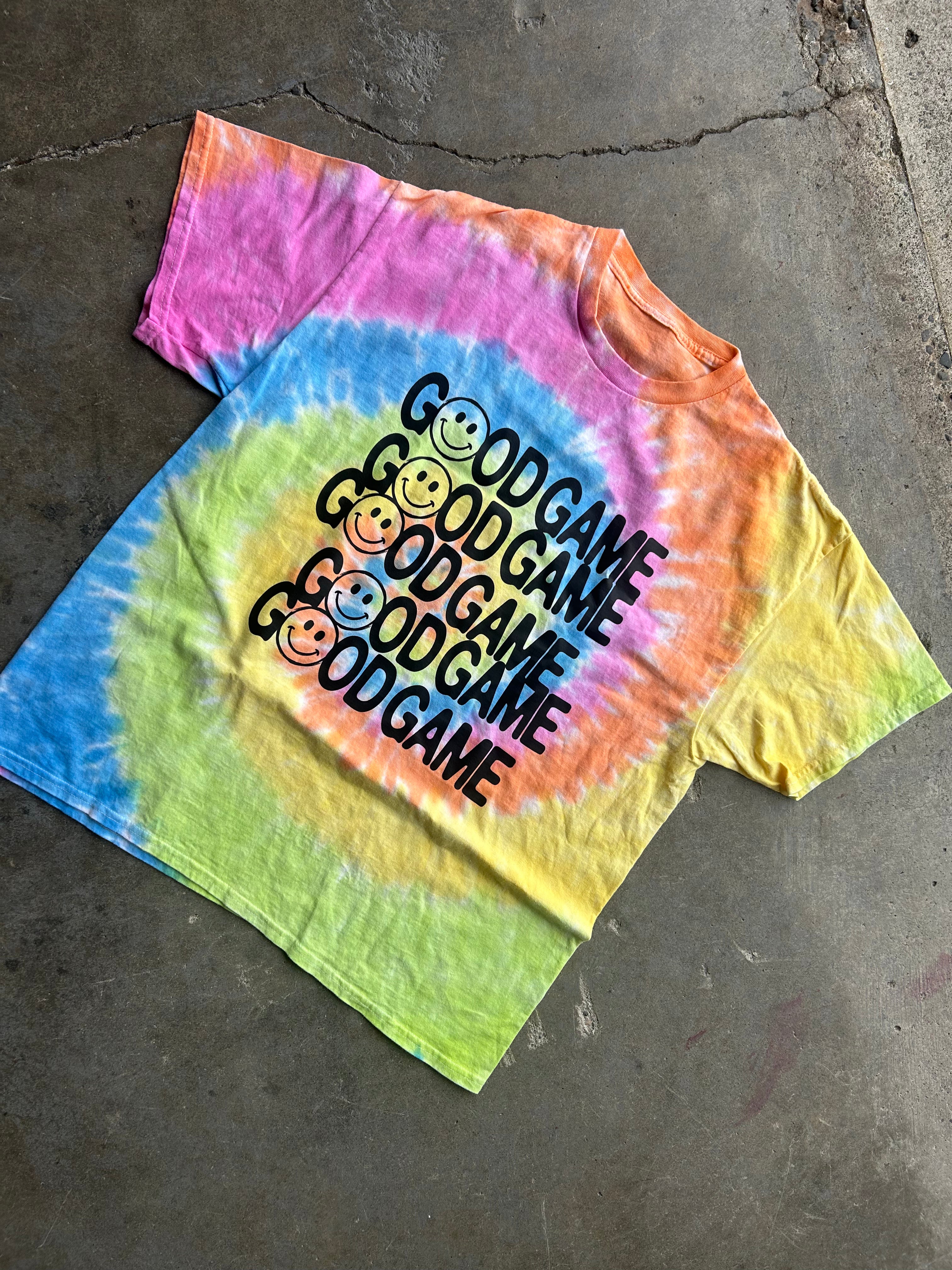 A short-sleeve tie-dye t-shirt laid flat on a concrete floor. The shirt features a spiral rainbow tie-dye pattern in pink, orange, yellow, green, and blue, with the text 'GOOD GAME' printed repeatedly in black over a stack of smiley-faced cartoon characters.