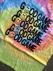 A close-up of a tie-dye t-shirt featuring the word 'GOODGAME' printed repeatedly in black, each accompanied by a smiley face. The shirt has a vibrant spiral tie-dye pattern in yellow, green, pink, blue, and orange.