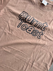 A close-up of a dusty rose or mauve t-shirt featuring the words 'Burnt Toast' printed in a dark navy outline font across the chest.
