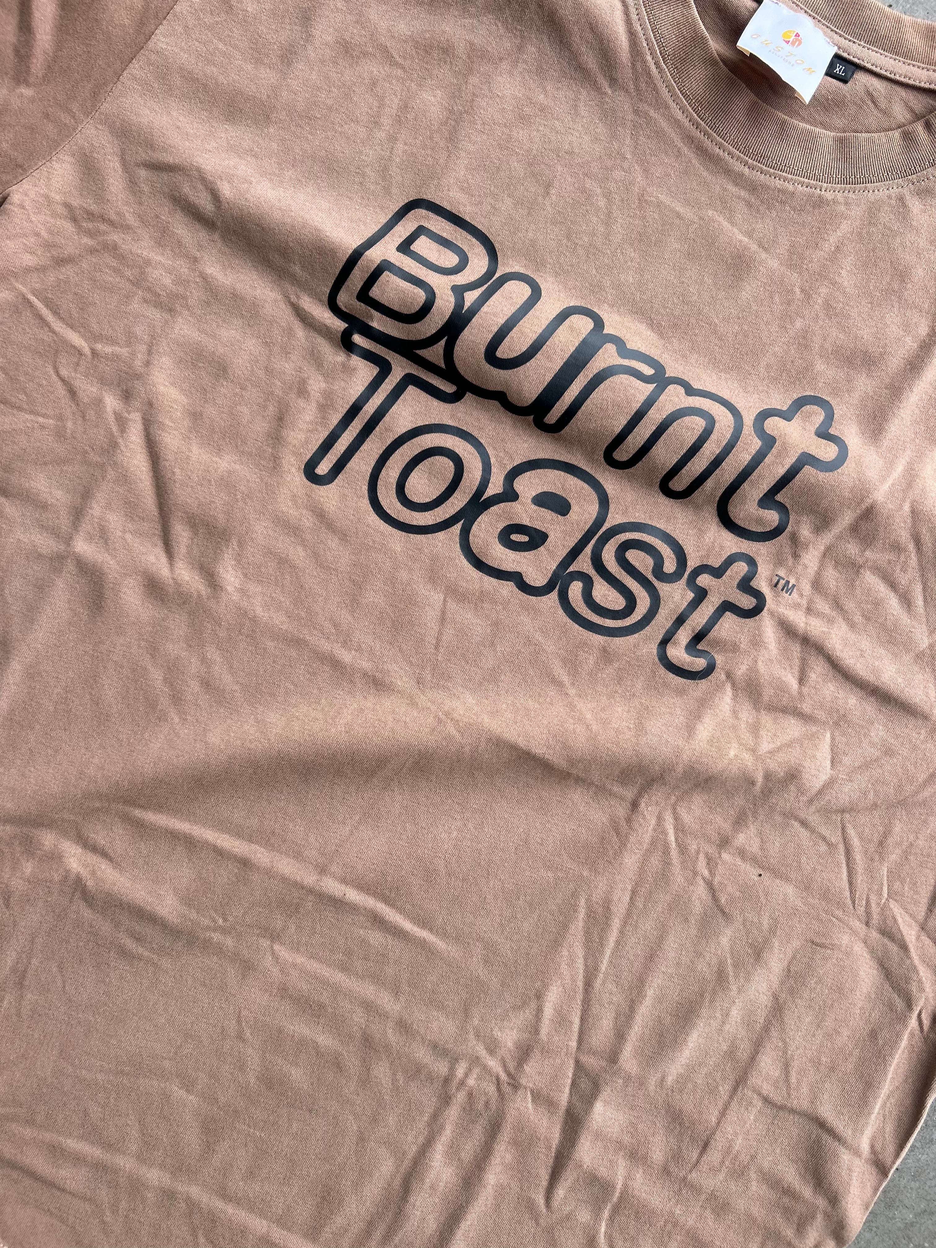 A close-up of a dusty rose or mauve t-shirt featuring the words 'Burnt Toast' printed in a dark navy outline font across the chest.