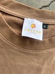 Close-up of a brown crewneck t-shirt showing a white brand label that reads 'CUSTOM SOLUTIONS' with a yellow and orange logo, and a black 'XL' size tag.