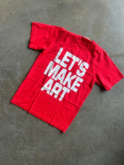 A red short-sleeve t-shirt with the phrase 'LET'S MAKE ART' printed in large white block letters, laid flat on a textured gray concrete floor.