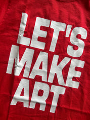A close-up of a red t-shirt featuring the phrase 'LET'S MAKE ART' printed in large, bold white block letters.