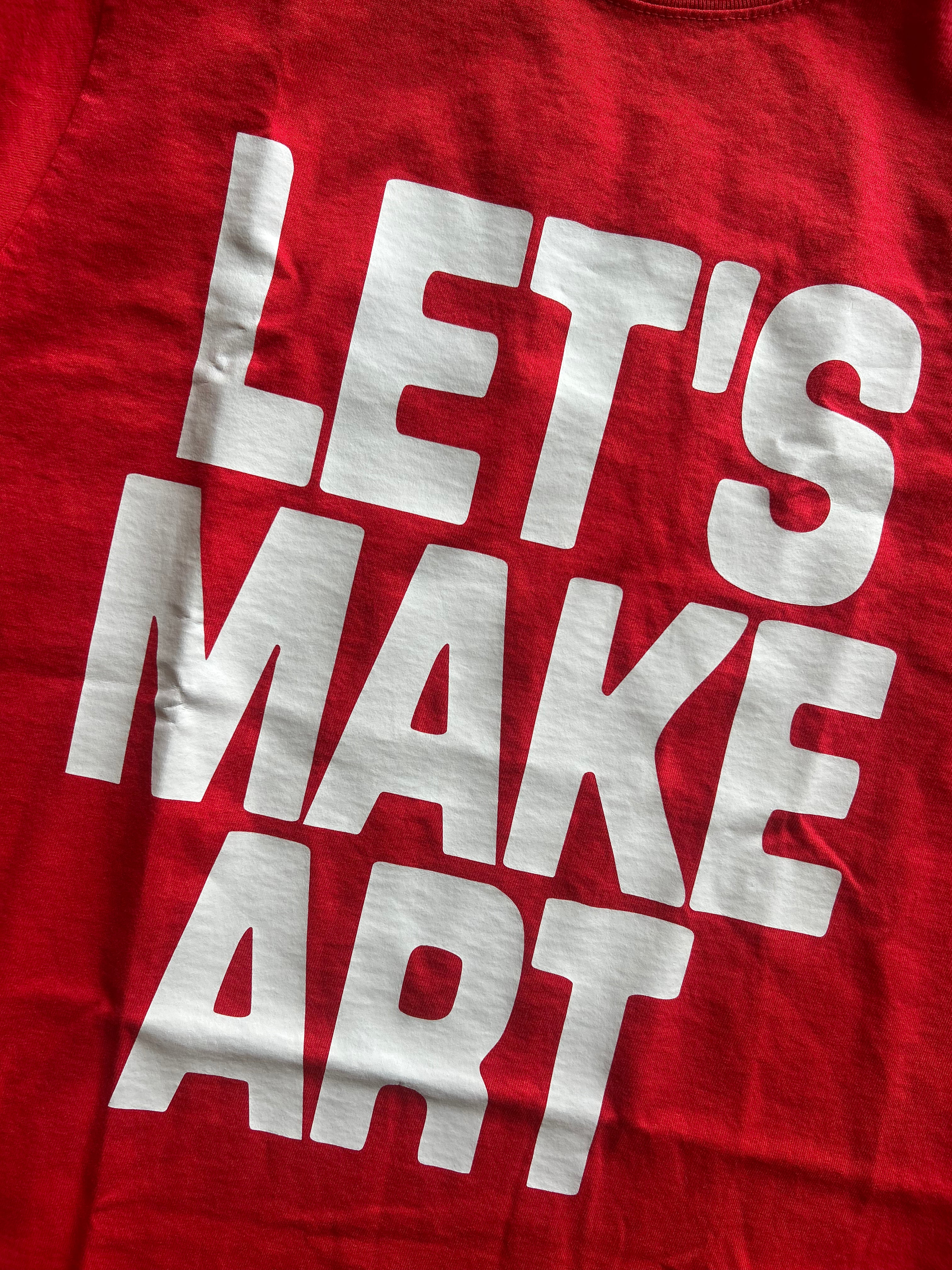 A close-up of a red t-shirt featuring the phrase 'LET'S MAKE ART' printed in large, bold white block letters.