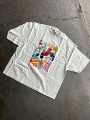 An oversized off-white cotton t-shirt with a colorful graphic collage on the front, laid flat on a textured concrete floor.