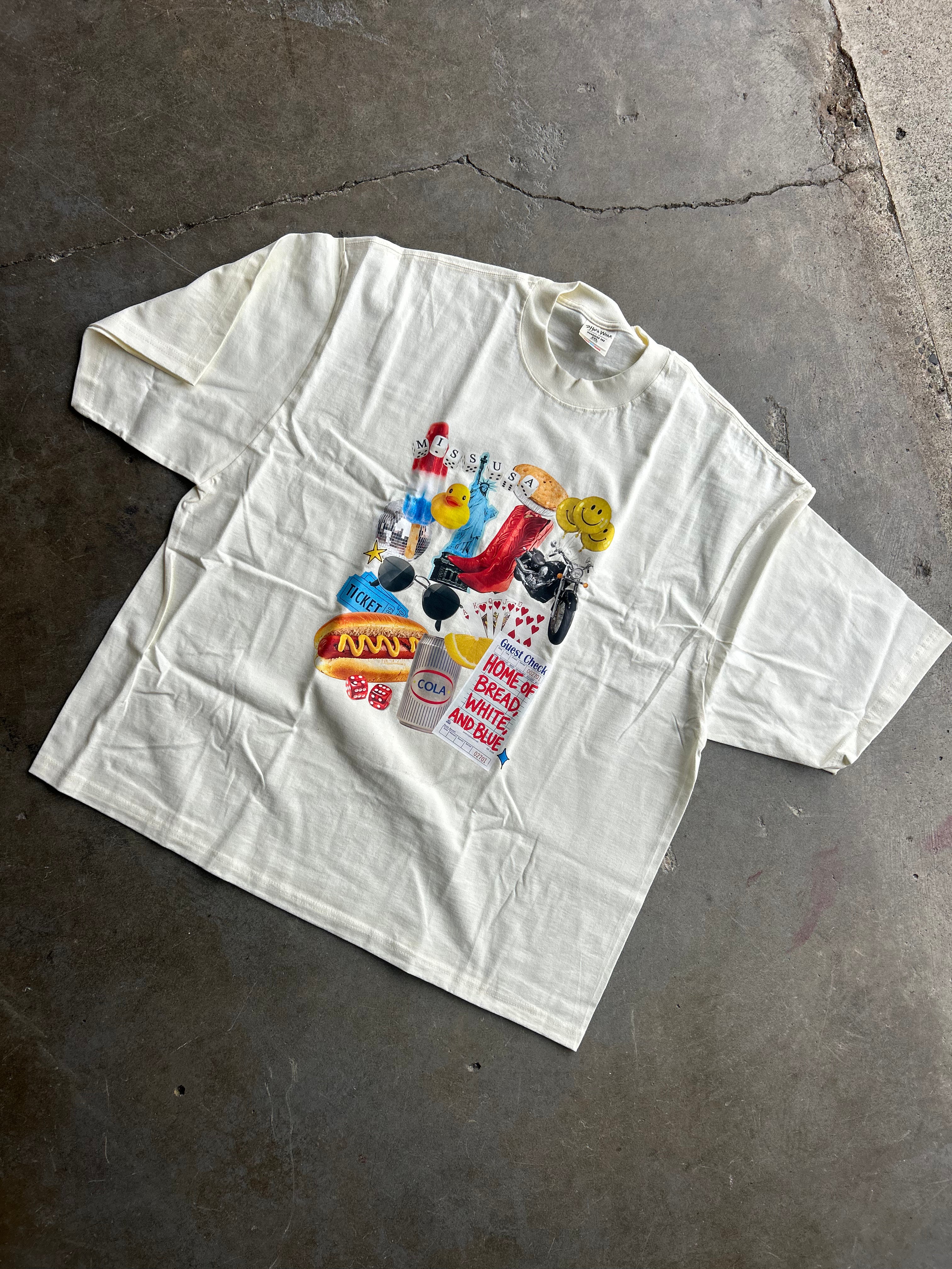 An oversized off-white cotton t-shirt with a colorful graphic collage on the front, laid flat on a textured concrete floor.