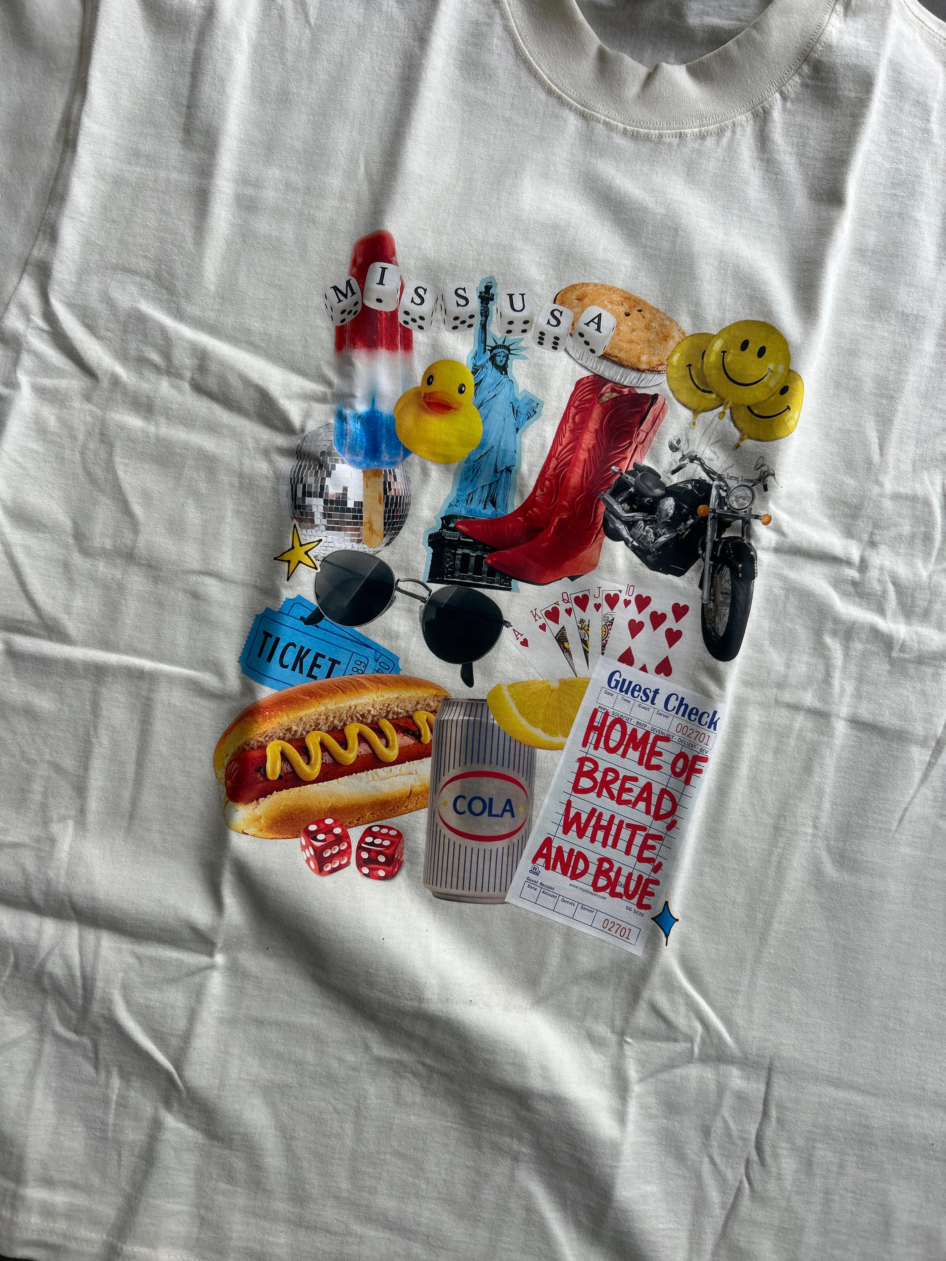 A white graphic t-shirt laid flat, featuring a colorful collage print on the front with various pop culture and social commentary elements including a hot dog, rubber duck, motorcycle, and a sign reading 'Home of Bread, White, and Blue'.