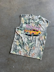 A sleeveless camouflage graphic t-shirt laid flat on a gray concrete floor. The shirt features a woodland camo pattern with a graphic of a yellow muscle car and the text 'Motherhood Never Dies' in pink and white.