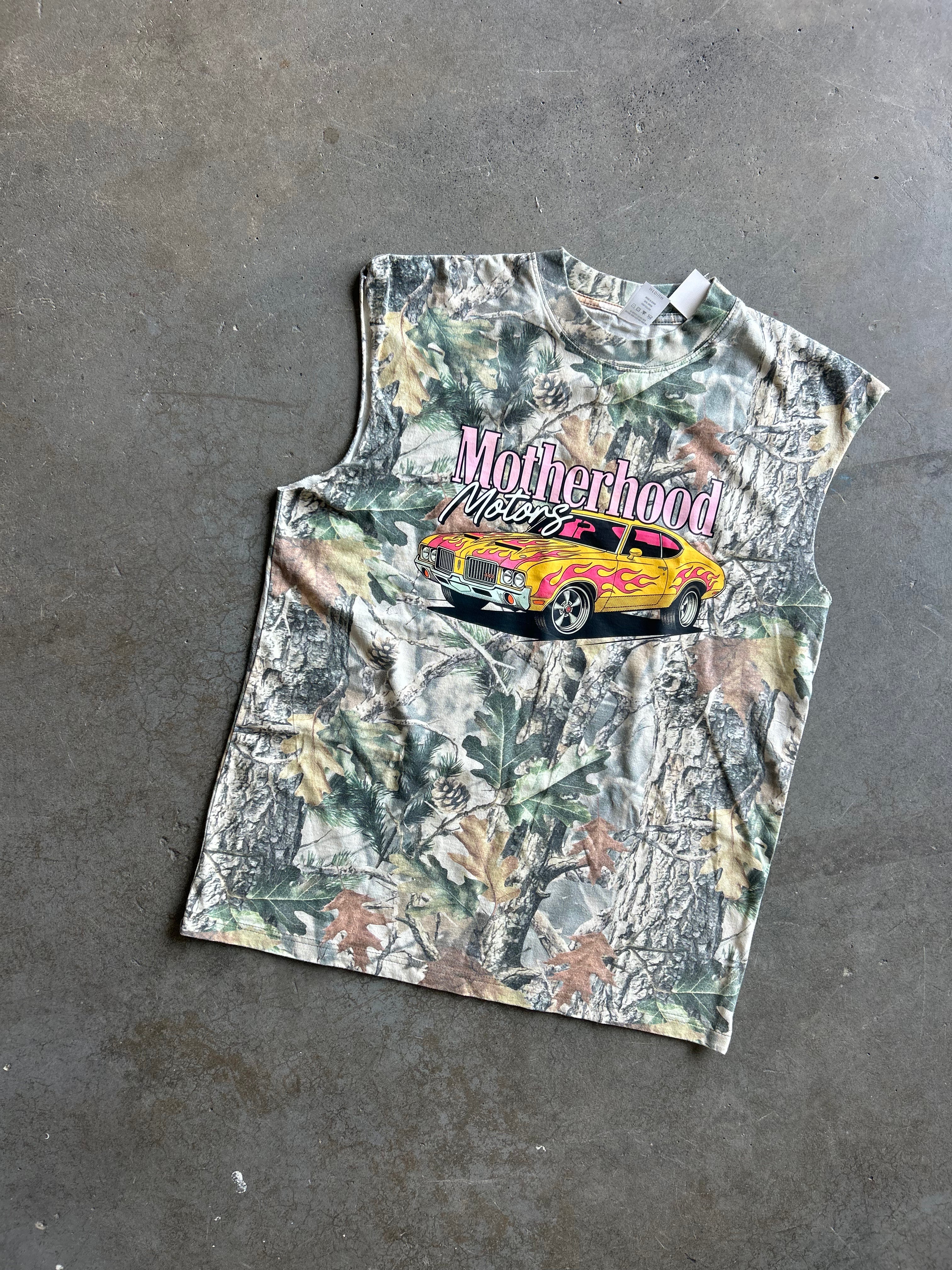 A sleeveless camouflage graphic t-shirt laid flat on a gray concrete floor. The shirt features a woodland camo pattern with a graphic of a yellow muscle car and the text 'Motherhood Never Dies' in pink and white.