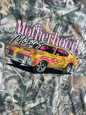 Close-up of a camouflage-patterned shirt featuring a large graphic of a yellow muscle car with pink flames and the text 'Motherhood Motors' in pink and white.