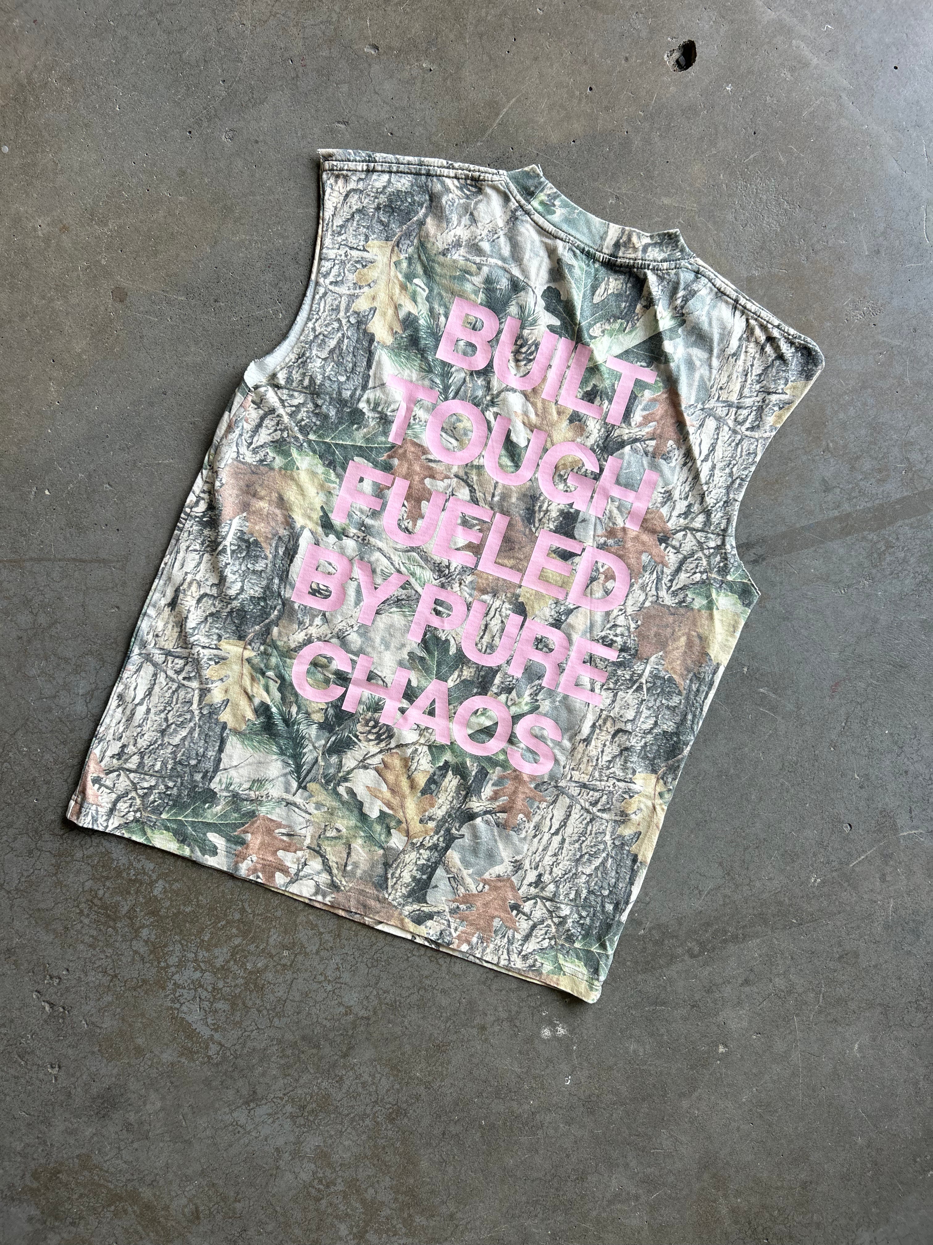 A sleeveless tank top with a woodland camouflage pattern and pink text reading 'BUILT TOUGH FUELED BY PURE CHAOS', laid flat on a gray concrete floor.