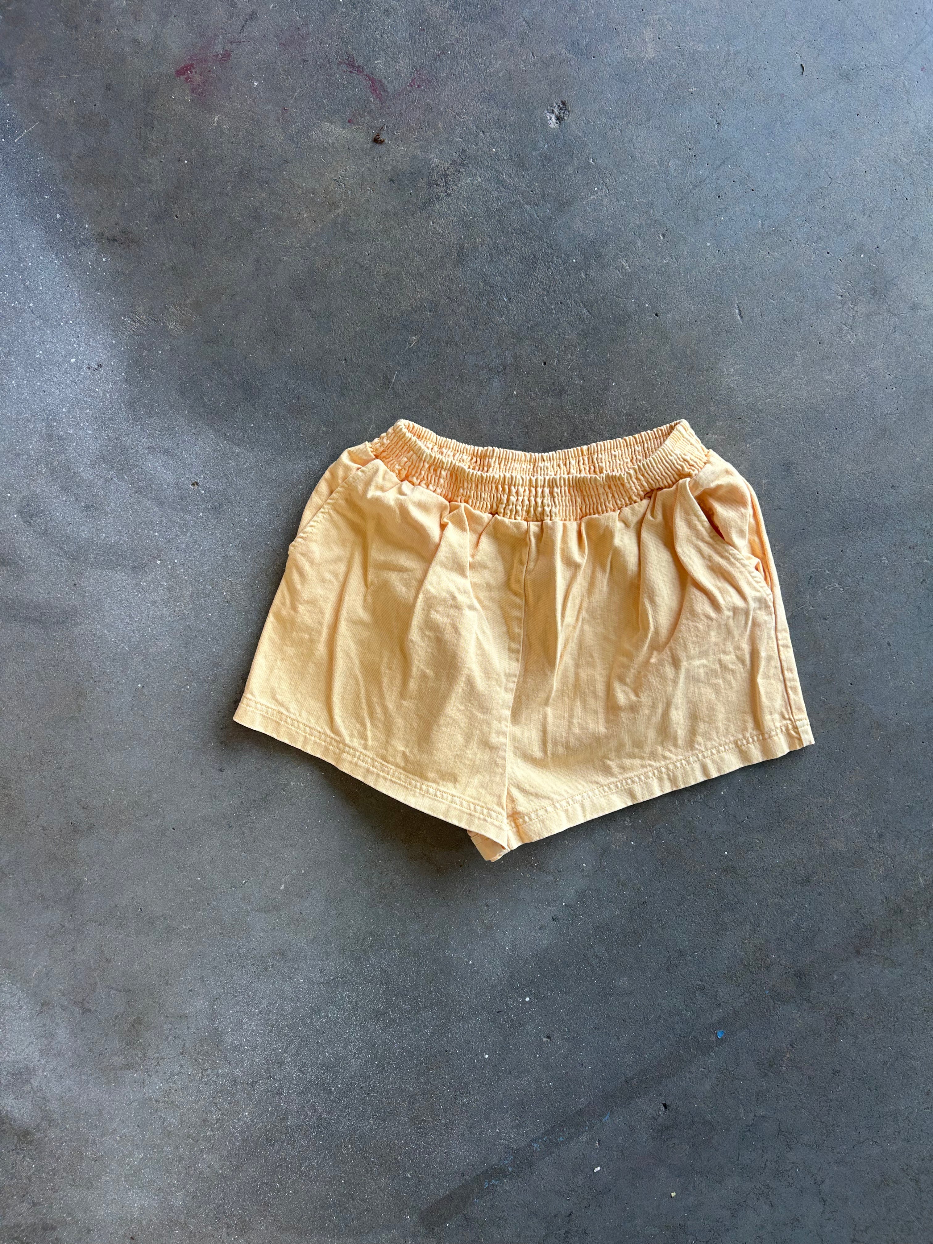 A pair of light peach or pale yellow shorts with an elastic waistband, laid flat on a textured gray concrete surface