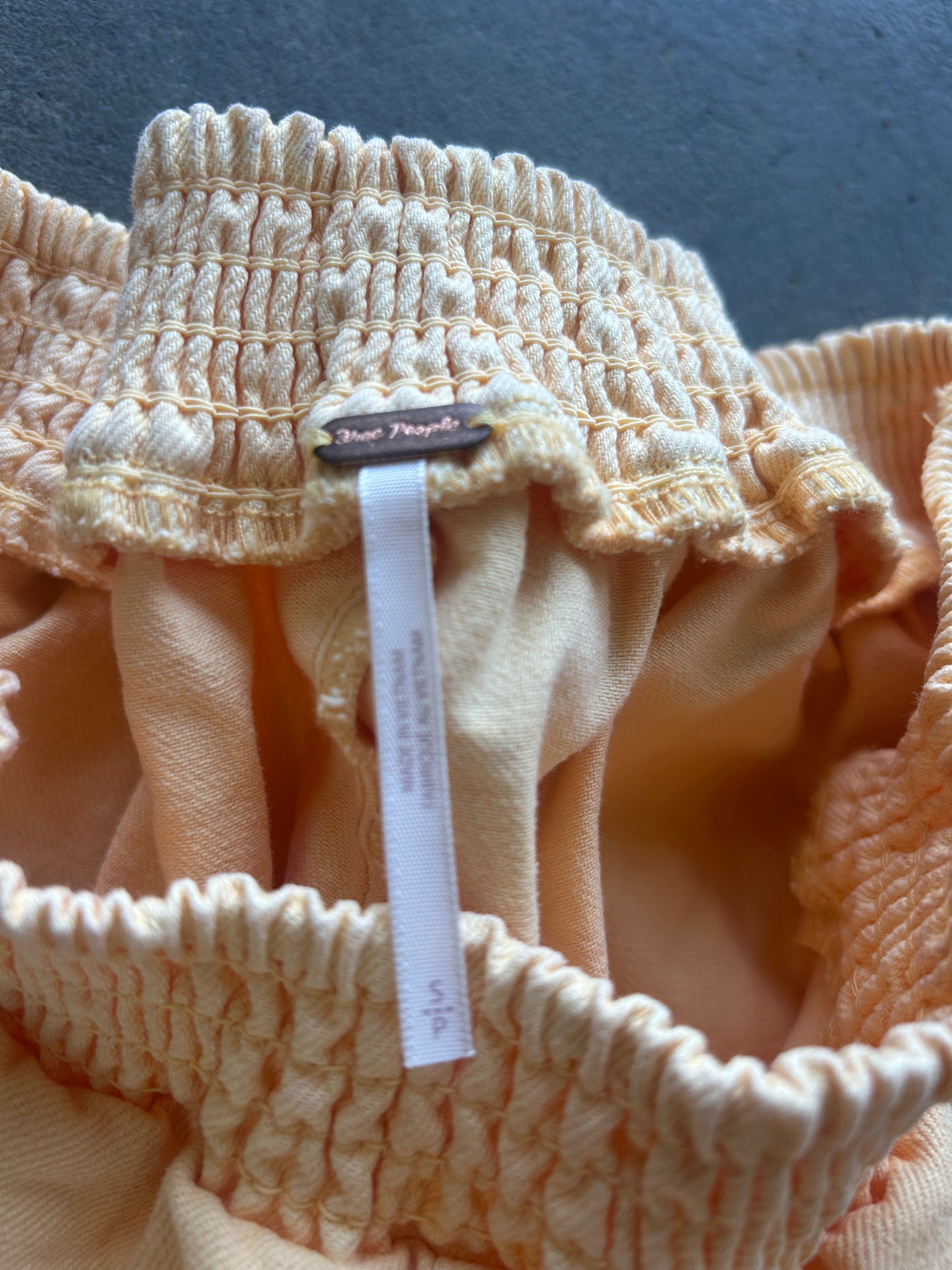 Close-up of a peach-colored smocked fabric garment, showing the elasticized ruffled waistband and a white fabric tag with a small brown label that reads Blue People