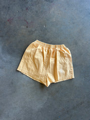A pair of light yellow linen shorts with an elastic waistband, laid flat on a textured gray concrete floor