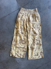 A pair of loose, cropped pants with an elastic waistband, featuring a beige and brown toile-style print, laid flat on a gray concrete floor