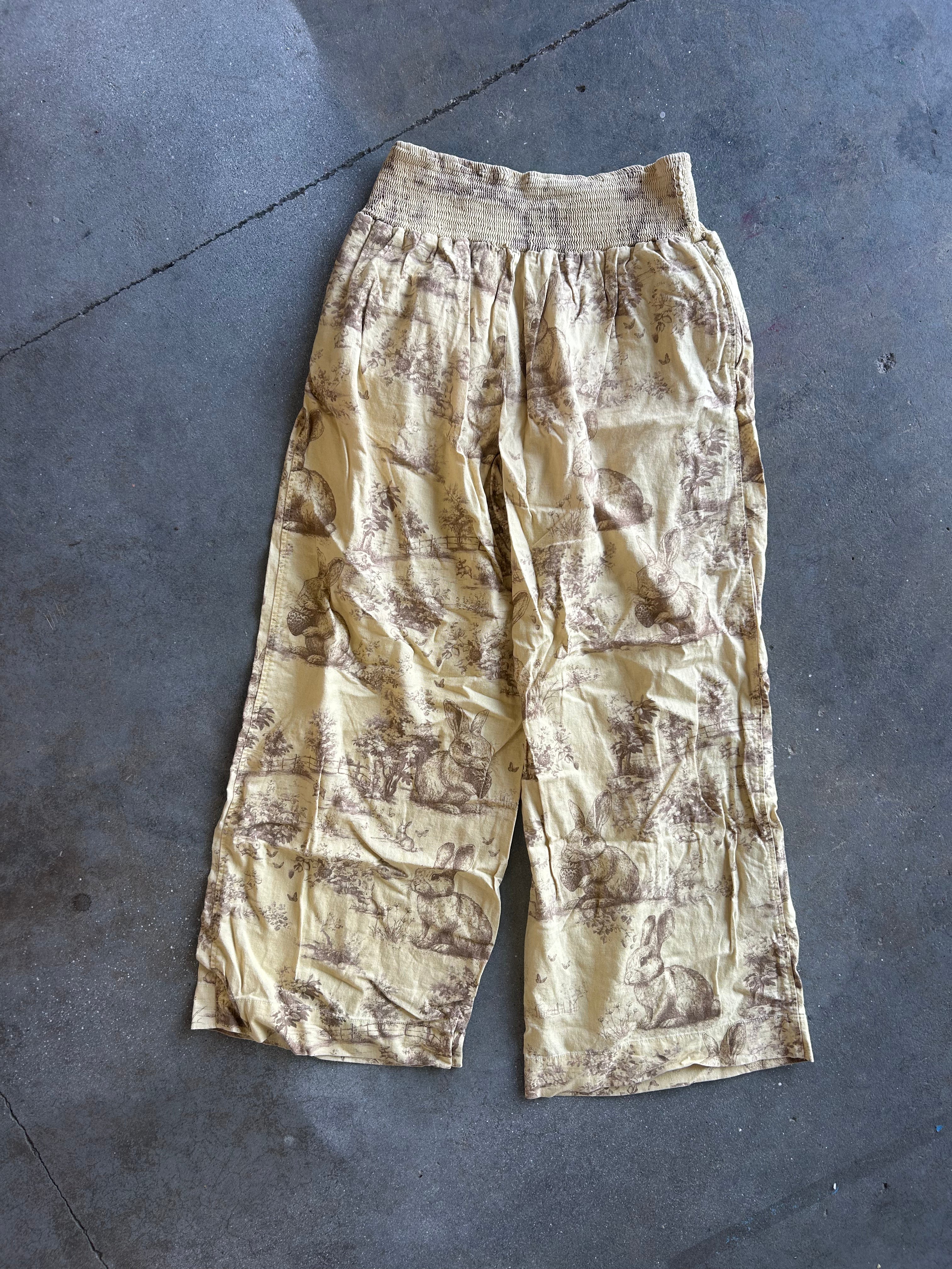 A pair of loose, cropped pants with an elastic waistband, featuring a beige and brown toile-style print, laid flat on a gray concrete floor