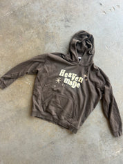 A dark brown washed hoodie with 'Heaven Made' in cream text and small starburst graphics on the chest, laid flat on a concrete floor